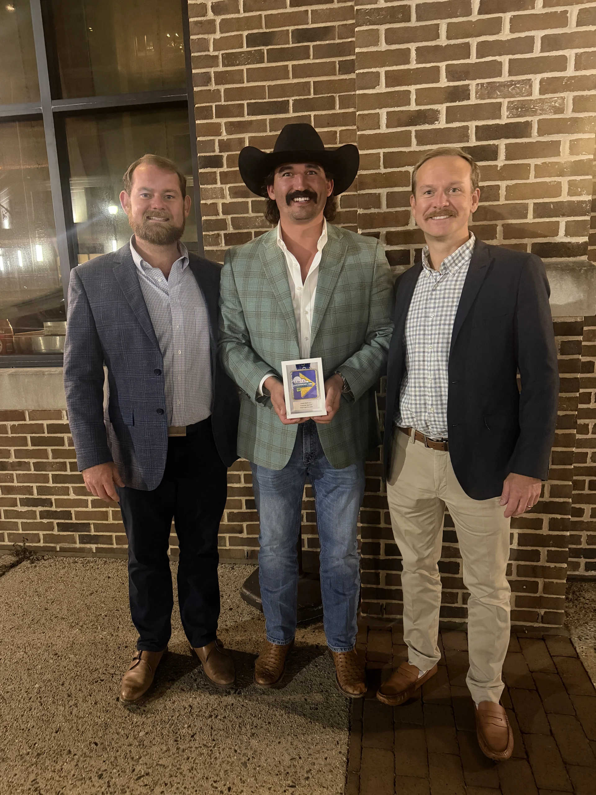 Three men in sport coats stand in front of a brick building at night, and the man in the center, wearing a black cowboy hat, holds a framed West Fight On medal with a plate reading Green Earth LLC, Supporter Sponsor, West Fight On 2026.