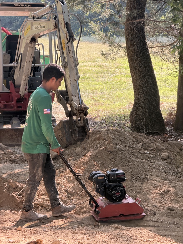 Green Earth crew member using a plate compactor near a tree
